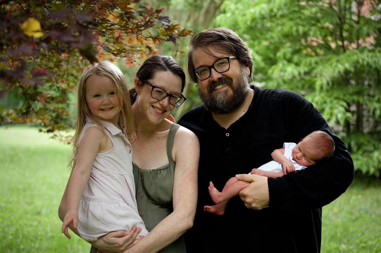 june 1 Emma Garton and TJ sit outdoors with their children, Bonnie and baby Jesse. Bonnie is held on Emma's hip, smiling at the camera, while TJ holds Jesse, who is wrapped in white. The family is surrounded by green grass and leafy trees.