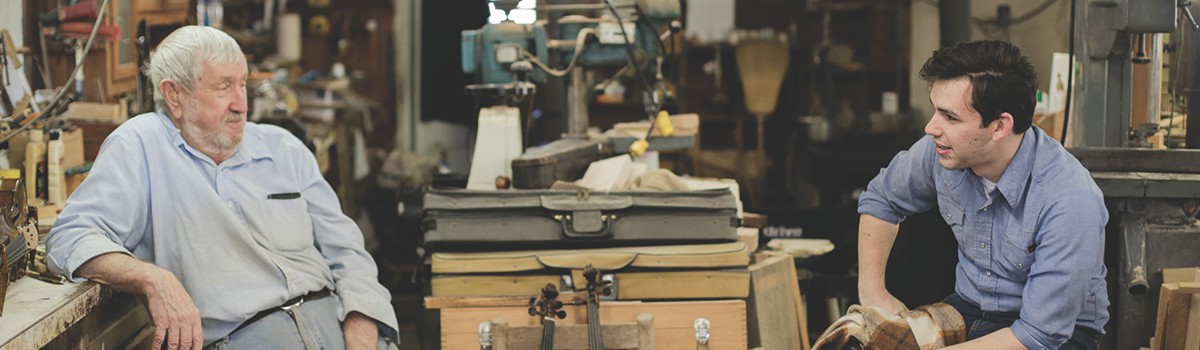 An older man and a younger man sit in a cluttered workshop, engaged in conversation. Both are wearing work shirts, and tools and equipment are visible in the background.