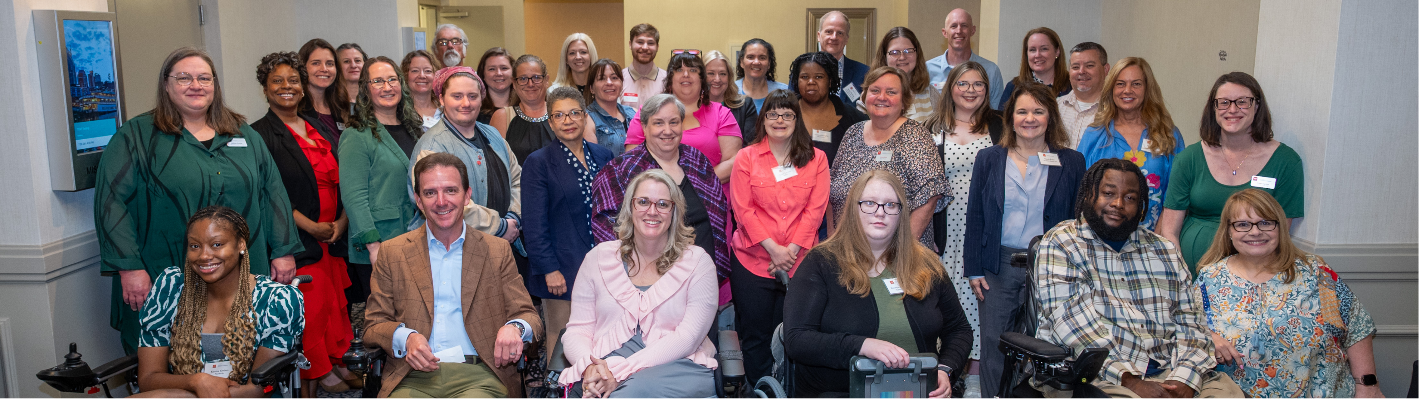 A group photo of the staff and members of the Tennessee Council on Developmental Disabilities. The diverse group consists of individuals of various ages, genders, and ethnicities, standing and sitting together in a well-lit conference room. Some individuals are using wheelchairs. They are dressed in a mix of professional and casual attire, with many smiling at the camera. The setting features neutral-colored walls, soft lighting, and a patterned carpet.
