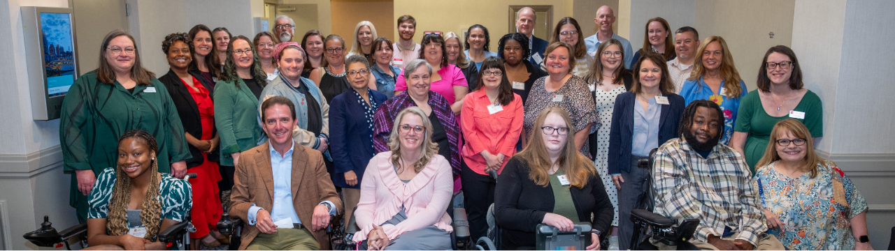 A group photo of the staff and members of the Tennessee Council on Developmental Disabilities. The diverse group consists of individuals of various ages, genders, and ethnicities, standing and sitting together in a well-lit conference room. Some individuals are using wheelchairs. They are dressed in a mix of professional and casual attire, with many smiling at the camera. The setting features neutral-colored walls, soft lighting, and a patterned carpet.