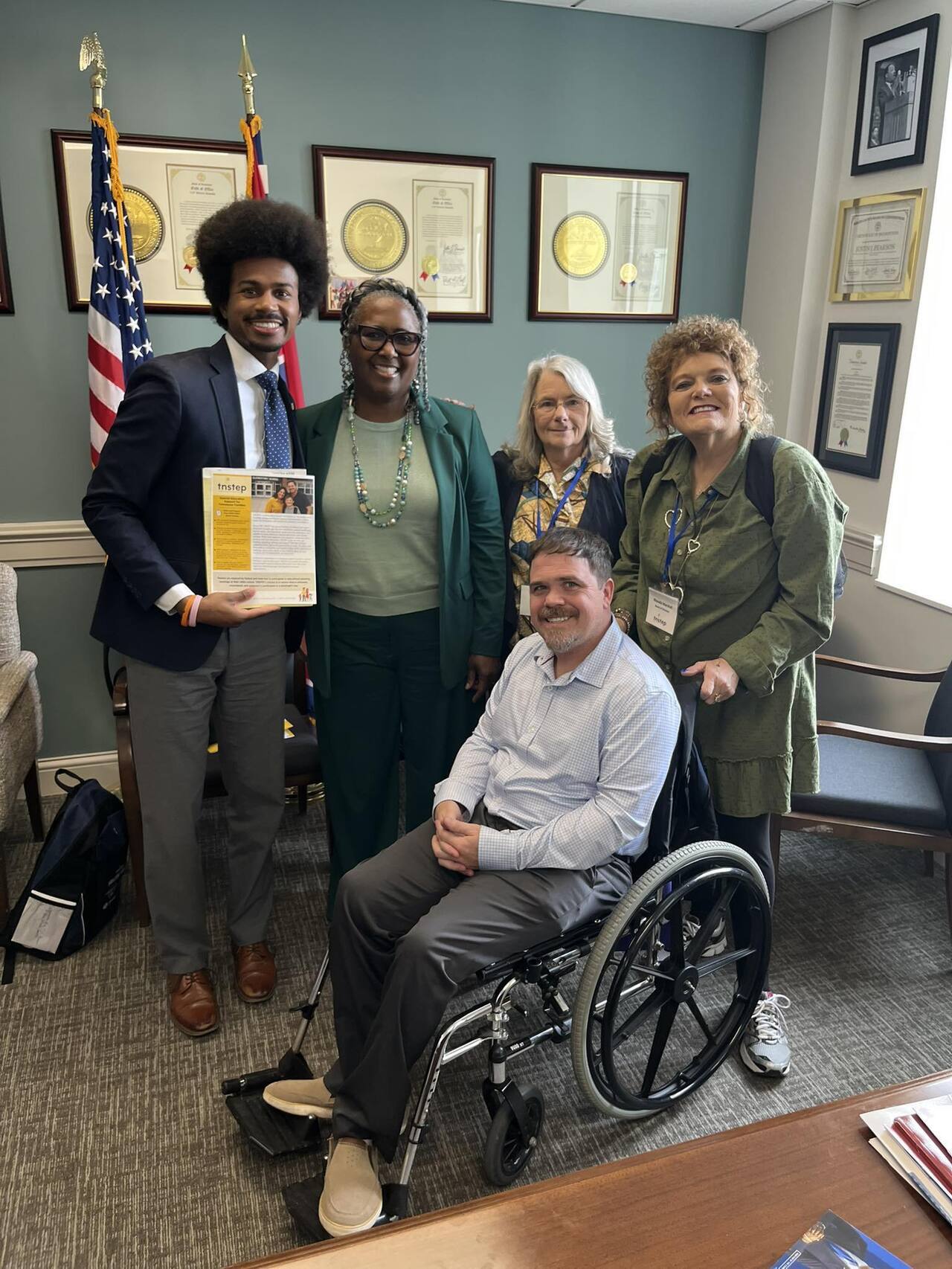 Five adults pose together in an office, smiling at the camera. From left to right: Justin Pearson, a dark-skinned man with a full afro wearing a suit and tie, holds a document; next to him stands a dark-skinned woman in a green suit with glasses; beside her is a light-skinned older woman with gray hair wearing a patterned scarf; on the far right is Rhonda Roberts, a light-skinned woman with short curly hair wearing a green dress. In front, a light-skinned man with short hair sits in a wheelchair, smiling. Framed certificates and flags are displayed on the wall behind them.