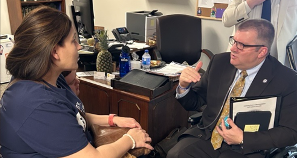 aith Henshaw, a woman with light skin and brown hair, wearing a navy blue shirt, sitting and attentively listening to Representative Michael Hale. Hale, a man with light skin, short-cropped hair, and glasses, is dressed in a dark suit with a gold-patterned tie. He is gesturing with his hand while speaking and holds a folder and a notepad. They are in an office setting with a wooden desk cluttered with papers, a pineapple, water bottles, and office supplies. A person in a white coat is partially visible in the background.
