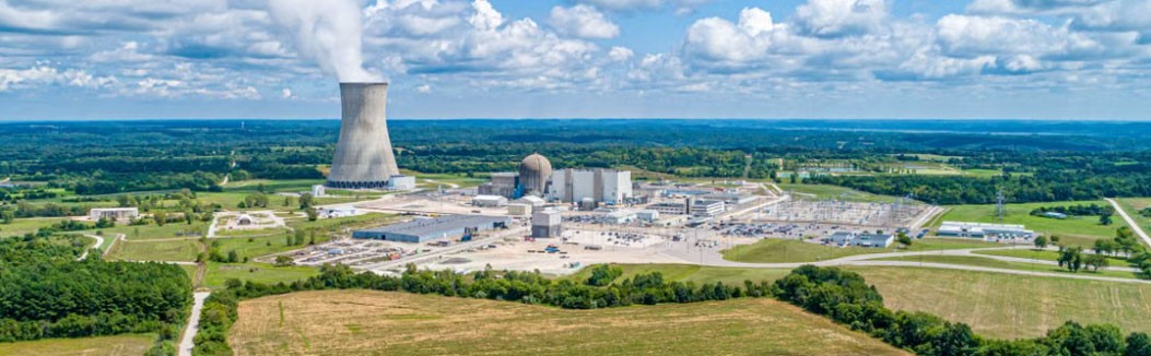 Aerial view of a power plant with a cooling tower releasing steam under a blue sky with scattered clouds.