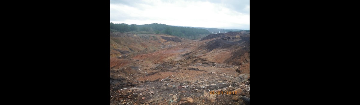 A wide mining pit stretches into the distance with reddish soil and scattered debris. Dark spoil piles rise along the right side of the pit. A forested ridge lines the horizon under an overcast sky. An orange date stamp reads 03/01/2017 near the lower right corner.
