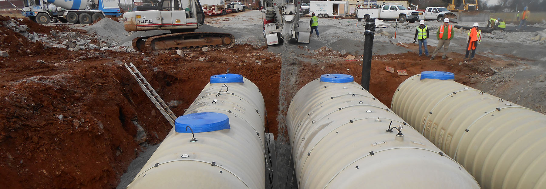 Three underground storage tanks being installed into a tank pit.  Tank pit being filled with gravel with workers surrounding the pit supervising.