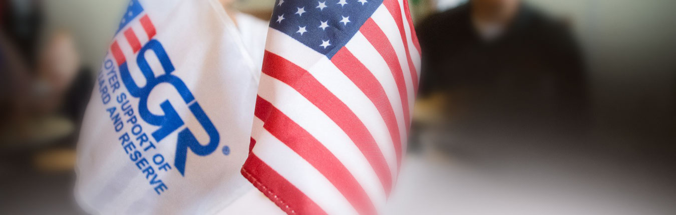 Two small flags sit on a table, one is the red, white and blue American Flag, and the other is a white flag with the ESGR, Employer Support of the Guard and Reserve logo, people are blurred and sit in the background at a meeting table.