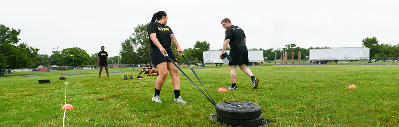 Photo of female Soldier in black tshirt with ARMY written in Yellow and black shorts pulling weights across green grass outside. 