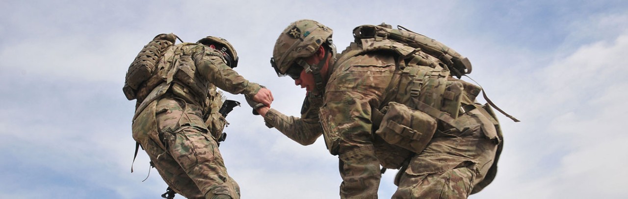 A Soldier in full combat uniform pulls another Soldier up off of the ground. A blue sky with clouds in the background.