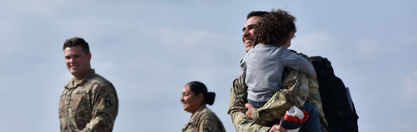 A Soldier picks up and hugs his child as arrives home from deployment. Other Soldiers in the background look on and walk to their awaiting families.