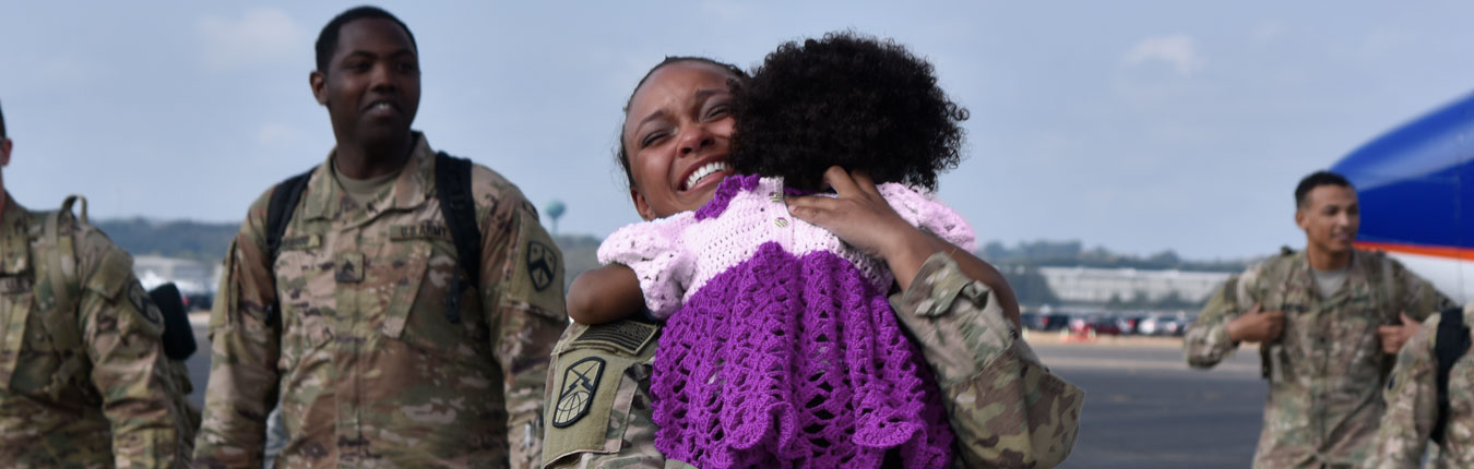A female Tennessee National Guard Soldier on an airport runway hugging her child after arriving back from deployment. Other Soldiers are in the background smiling and walking away from the plane.
