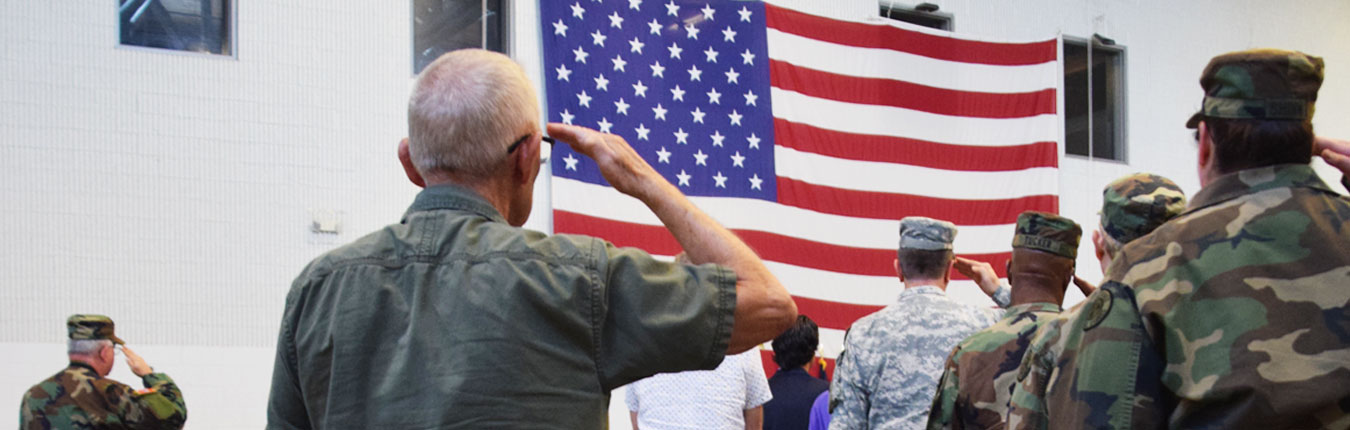 An older man saluting at a large American Flag hanging on the wall inside a military armory. Other Soldiers of different organizations are also at attention and saluting the flag.