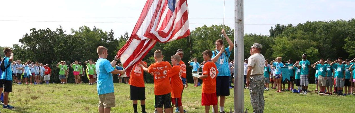 Children at a Tennessee National Guard Youth Program Event - Children in bright colored shirts along with a Tennessee National Guard Soldier help to raise a large American Flag outside, while other children in brightly colored shirts salute.