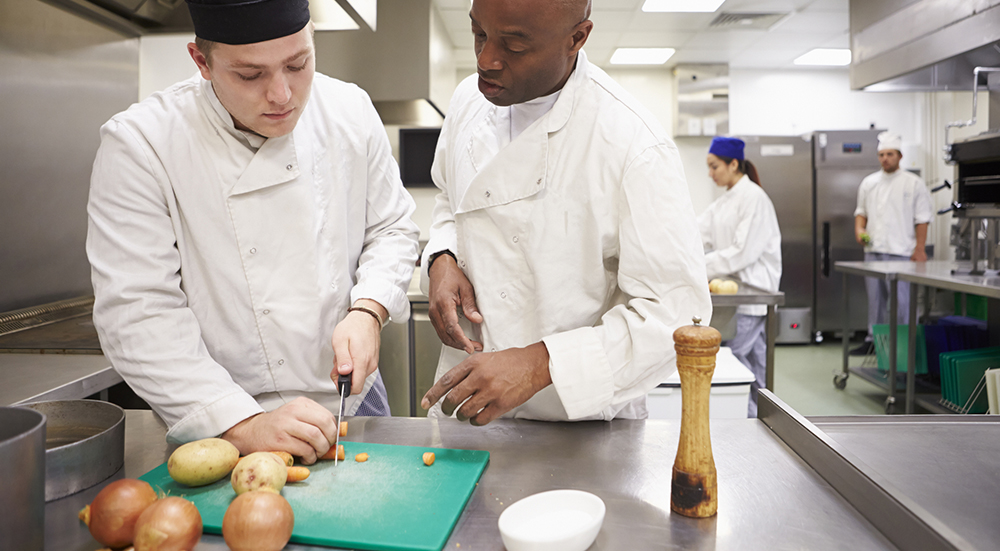 A chef supervisor watches over a young chef cutting vegetables