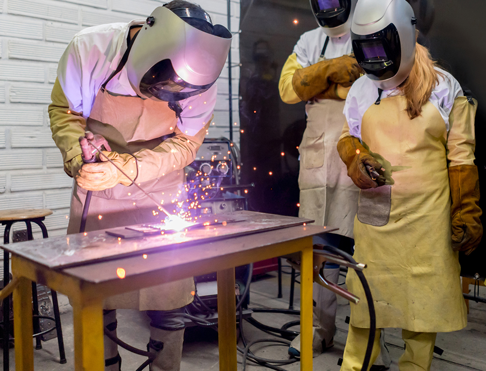 Three people wear helmets, protective suits and goggles while one person uses a welding torch