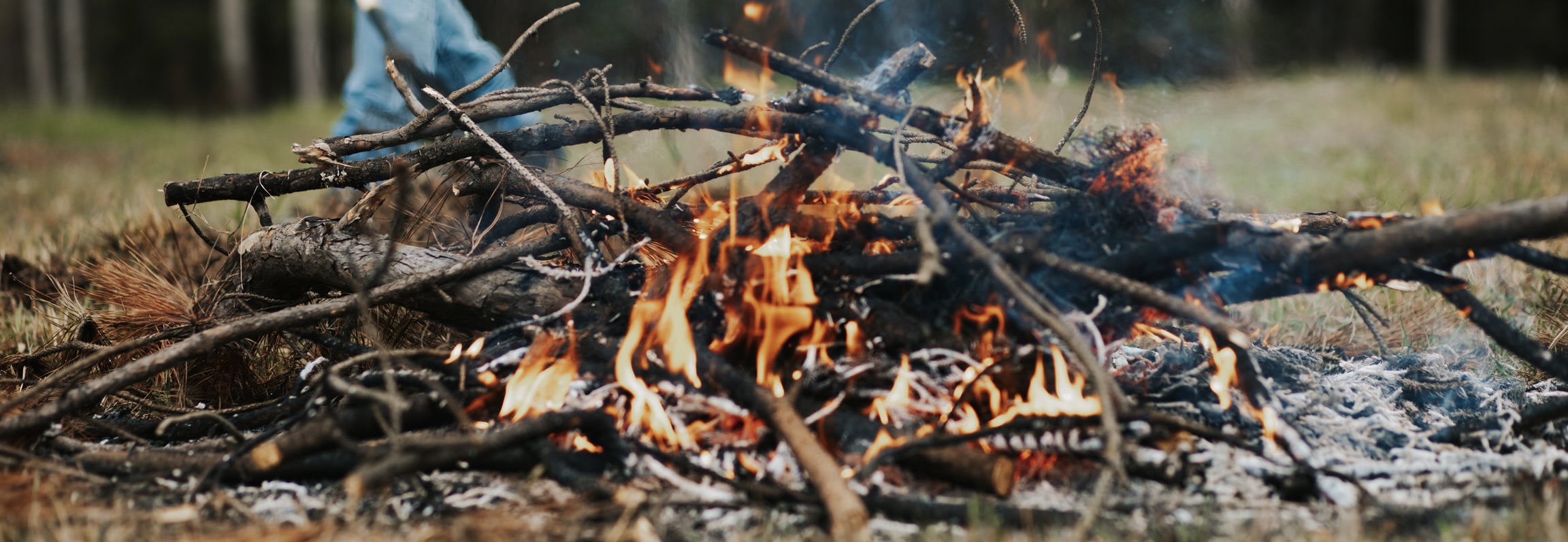 A pile of burning branches and logs with visible flames and smoke, set on a grassy forest floor, with a person’s legs in blue jeans partially visible on the left side.