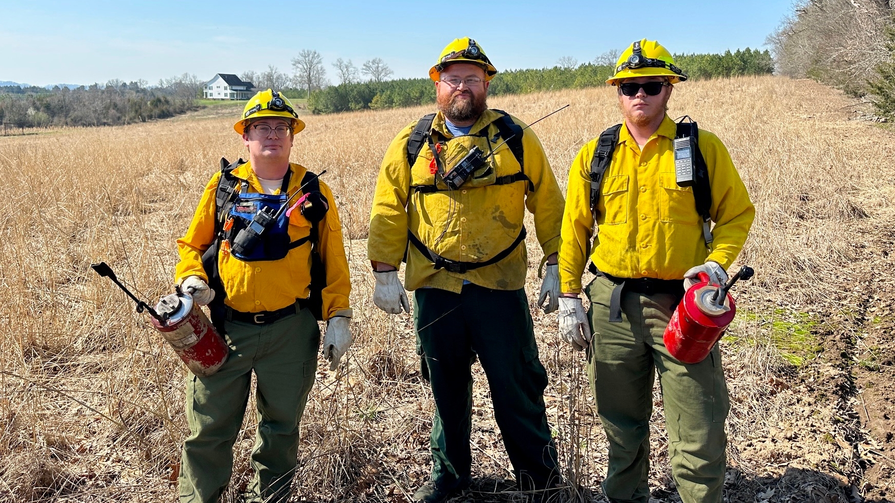 Three wildland firefighters in yellow helmets and jackets, green pants, holding red extinguishers, stand in a dry grassy field with a house in the background.