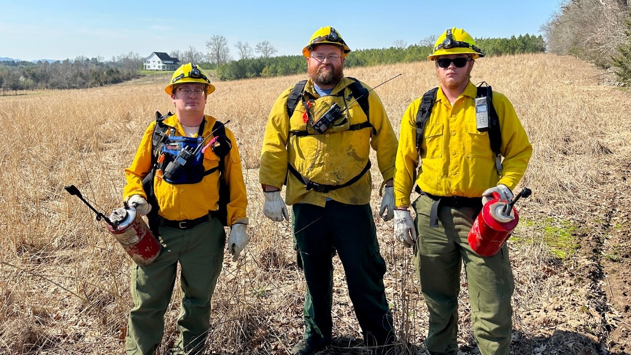 Three wildland firefighters in yellow helmets and jackets, green pants, holding red extinguishers, stand in a dry grassy field with a house in the background.