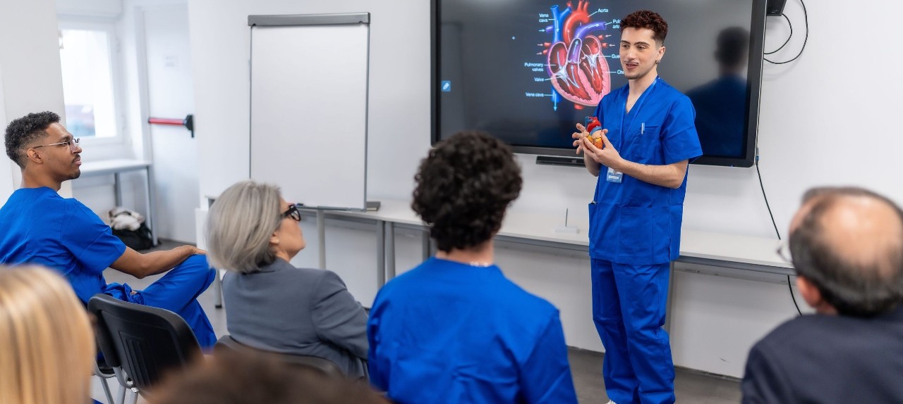 Man in scrubs talks in front of a classroom with an image of a heart on a screen behind him