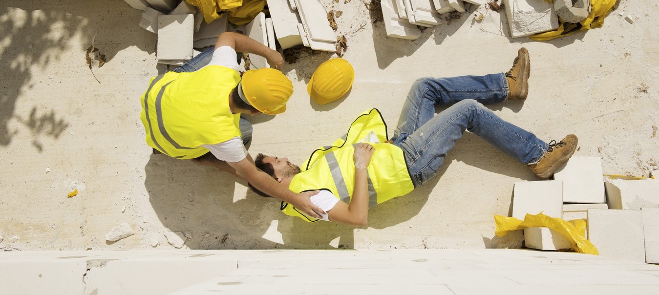 A construction worker wearing a yellow safety vest and hard hat kneels beside a fallen coworker on a construction site. The injured worker is lying on the ground among scattered white bricks and debris, while the other worker supports him and checks his condition. Both are wearing jeans, safety boots, and reflective vests.