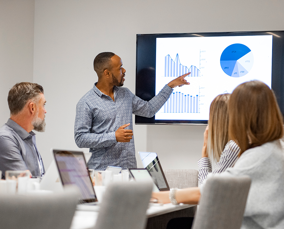 A man points out a graphic on a screen to people seated at a conference table