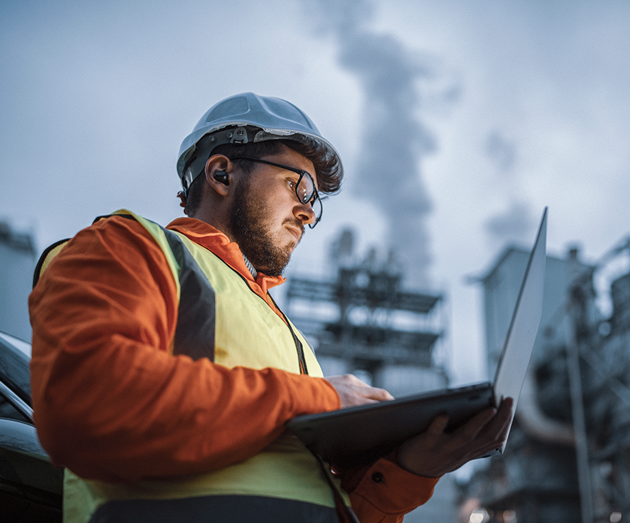 Man wearing a construction helmet looks at laptop at industrial site