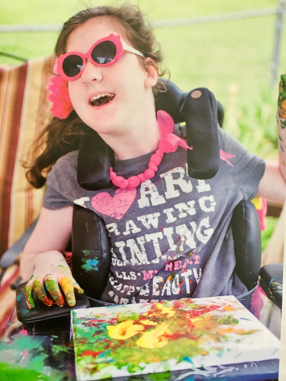 The photo shows a young girl with curly brown hair in a ponytail, with bright pink sunglasses and a big grin on her face as she sits outside in her wheelchair in the sunshine. Her hands and shirt are covered in paint as she creates a painting on a canvas resting on her lap