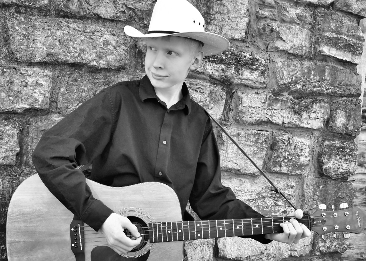 The photo of Andrew in this article is a black and white photo of a young man with light blond hair, a black button up shirt and jeans, and a cowboy hat. It looks like a professional headshot of a musician – he is leaning against a stone wall looking off to the left past the camera, smiling slightly and holding a guitar as though he is about to play.