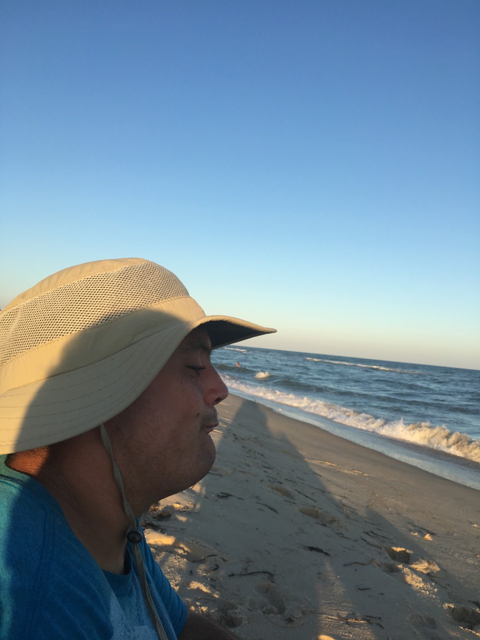 A close-up photo of a man with Down syndrome wearing a fisherman’s hat. He is at the beach, near the edge of the water, and the photo shows him on a sunny, windy day with the shadow of the photographer on the sand behind him.