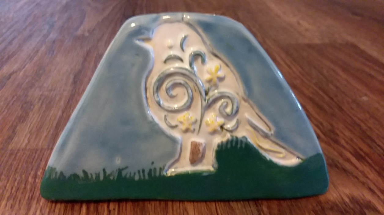 A photo of a piece of pottery that is shaped like a trapezoid, wider at the bottom and narrower on top. The bottom of the piece is painted green like grass. There is a detailed outline of a bird, a dove, perched on the grass with decorative swirls inside its outline. The rest of the pottery is gray, like a cloudy day.