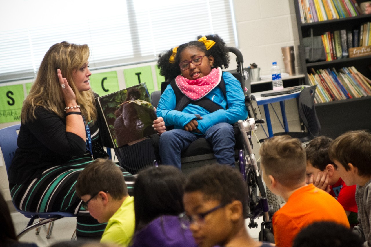 a teacher with blond hair sitting down in a classroom while she puts a hand up to her ear, as if to tell her students to speak up – she holds a book showing a photograph of a gorilla in a jungle. Next to her is Charlotte Bynum, Alison’s daughter, who is a young girl with pig tails and bright yellow scrunchies in her black hair, glasses, a colorful pink handkerchief around her neck and a smile as she sits in her wheelchair. Caption: Charlotte Bynum’s teacher interacts with her as they read together with her class