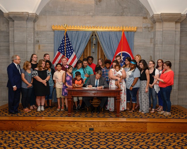 A couple of dozen children and adults stand behind and surrounding Governor Bill Lee who sits at a table and signs a bill creating the Deaf Mentor Program at a bill signing ceremony; caption reads: Advocates surround Governor Bill Lee as he signs the Deaf Mentor Pilot Project bill into law in 2019