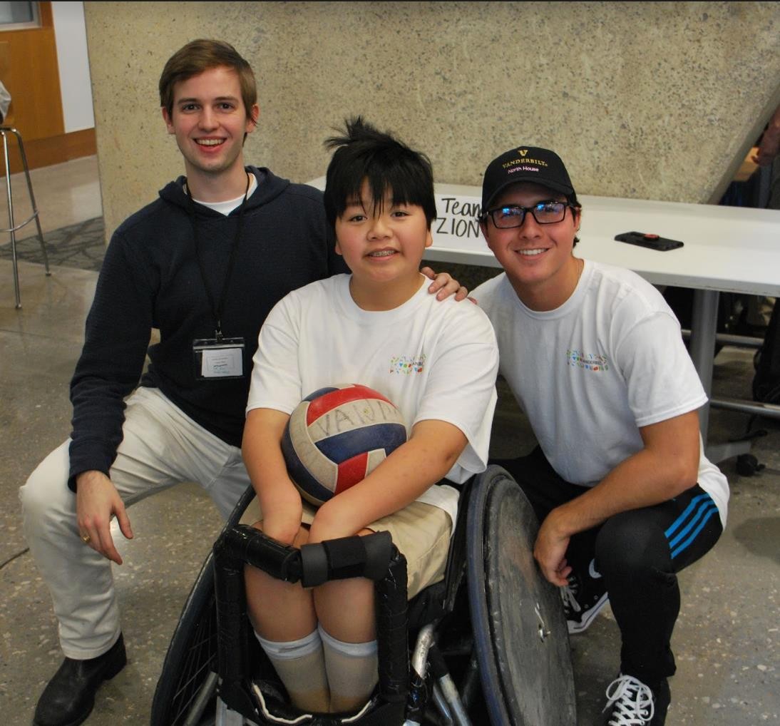 Two college students are shown with a pre-teen or young teenager with a disability who is a wheelchair user