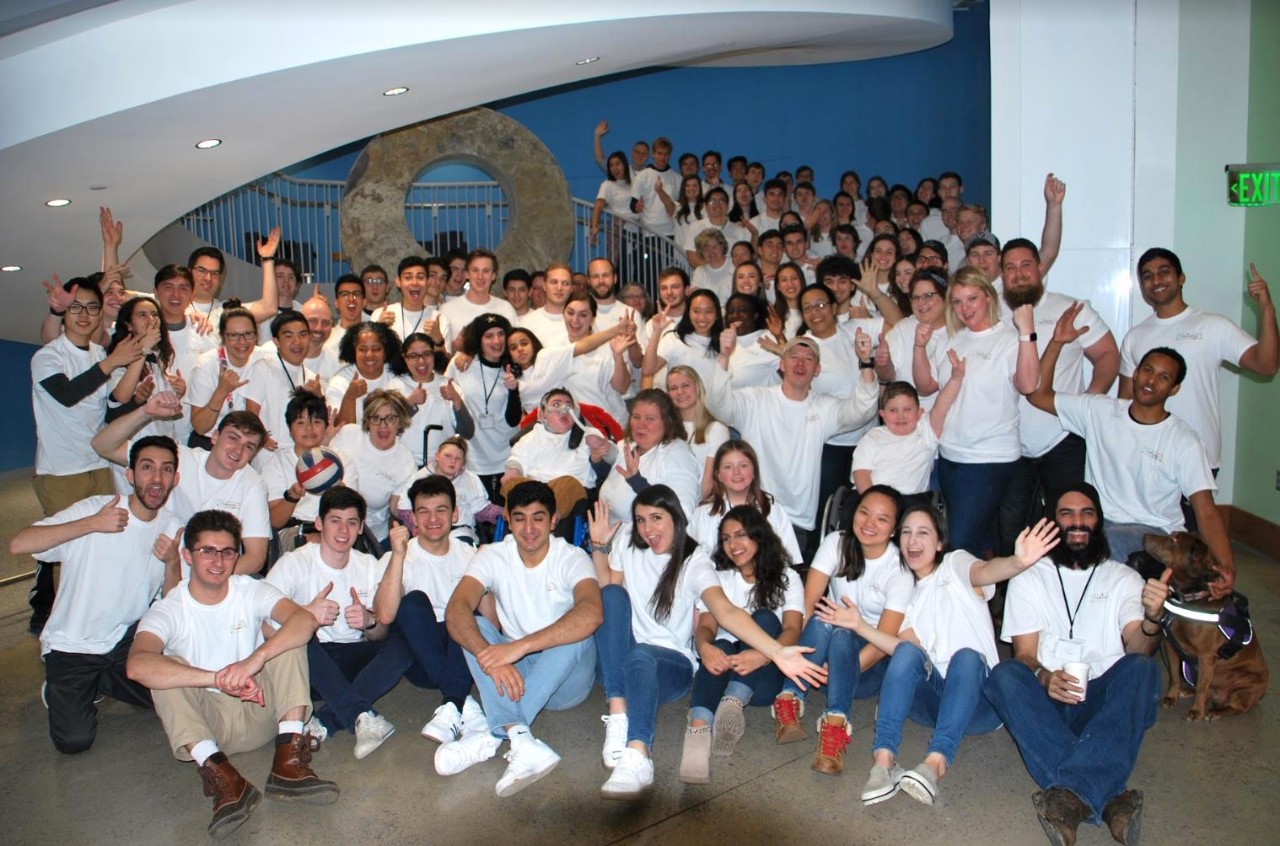 a large group photo of maybe 3 dozen people of all ages, all wearing matching white T-shirts, sitting and standing together, smiling and many are raising their hands in celebration. The group includes many college students, and children and adults with various types of disabilities and their families