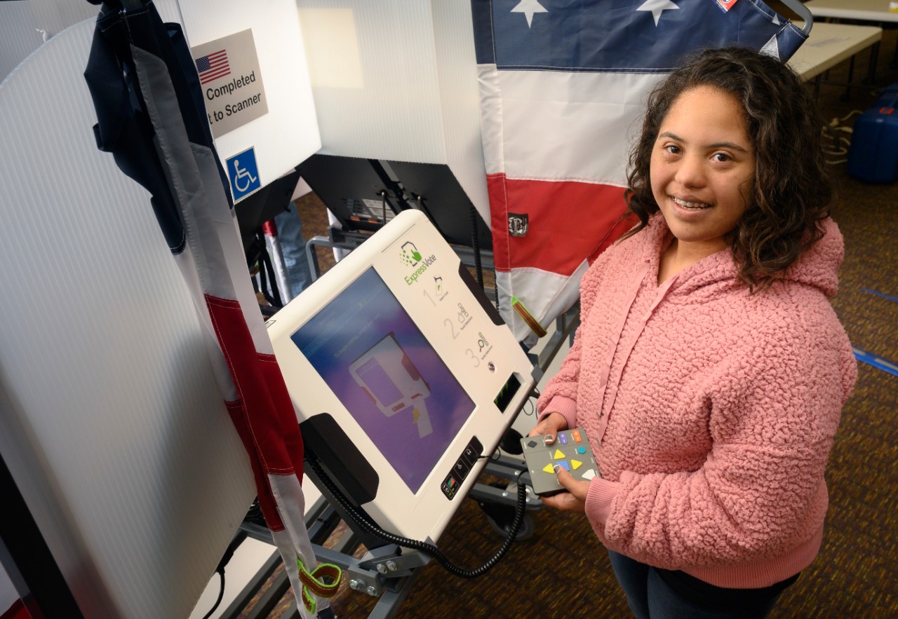 A young woman with Down syndrome and brown curly hair wearing a pink fluffy pullover sweater stands at a voting machine holding the keypad to navigate the machine to cast her ballot; she smiles at the camera