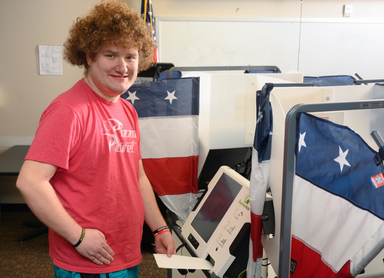 A young man named Zach with autism is wearing a bright red shirt and posing by a voting machine bookended with American flags; he’s looking at the camera and placing a mock ballot into the machine