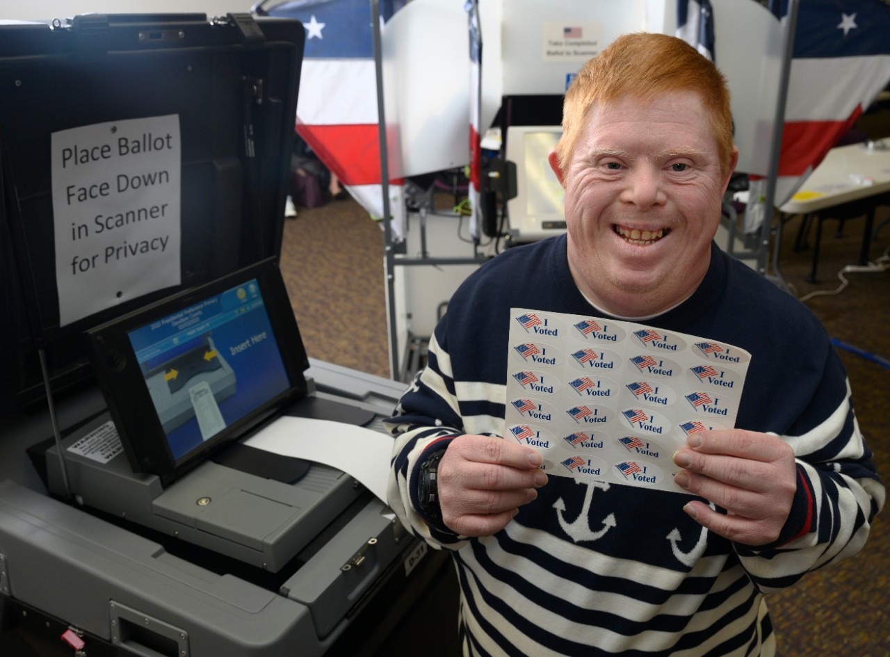 A middle-aged man with Down syndrome, red hair and a big smile holds up a sheet of “I Voted” stickers, standing by a voting machine and smiling at the camera.