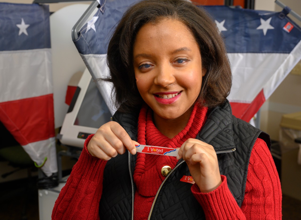 a photo of a young black woman with short brown hair, a bright red sweater with a black vest over it and a lovely smile holding up an “I voted” sticker in front of her, showing it to the camera. Behind her are voting machines with American flag banners nearby. 