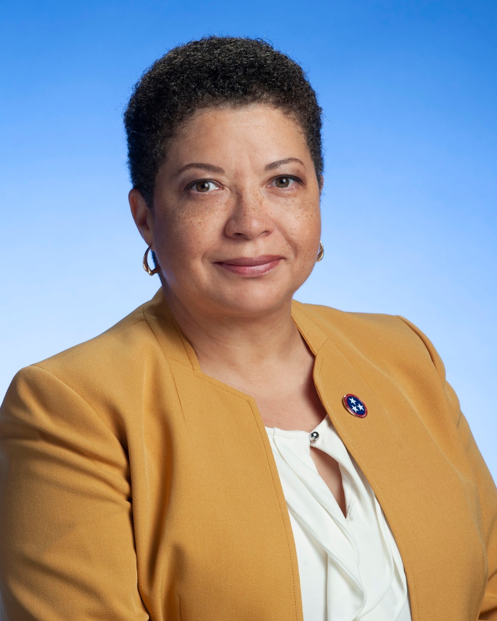 Cathlyn’s headshot. She is an African American woman in a dark yellow blazer with a state of Tennessee pin on her lapel, small hoop earrings, short hair and a kind smile.