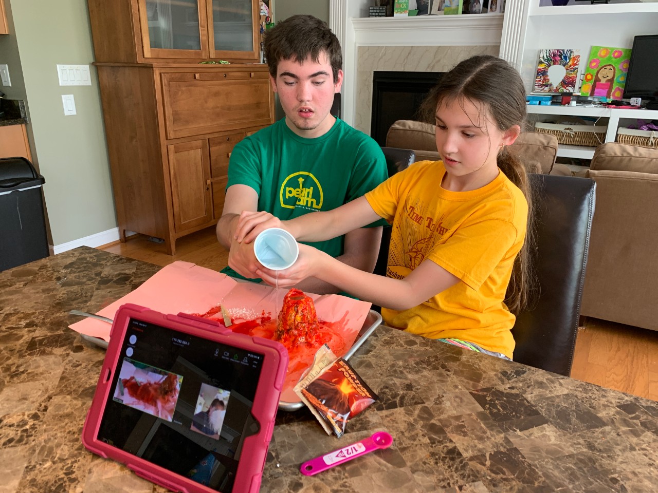 The two siblings sit at a kitchen counter and do a science experiment with a model volcano.