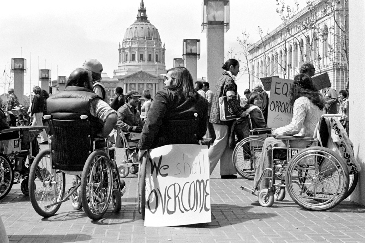 photo is in black and white and shows a number of people with visible disabilities, including people of all ages using wheelchairs, at a protest with a large domed federal building behind them in the background of the photo. A couple of protest signs on posters are visible in the crowd – the one most prominent in the photo reads “We Shall Overcome” – it’s attached to the back of a wheelchair where a man with long brown hair is seated, talking to other protesters
