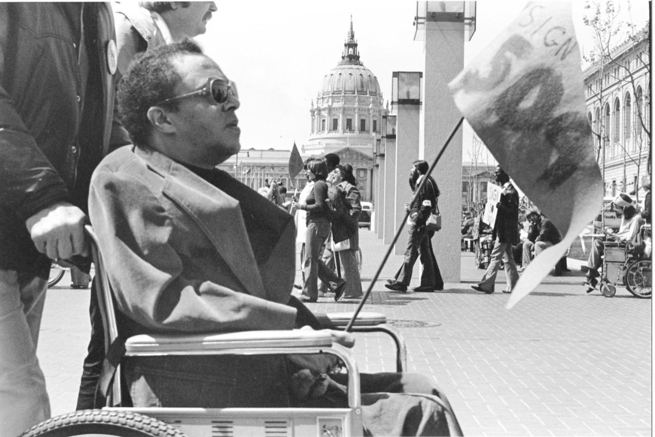 a close-up black and white photo of a man in a wheelchair being pushed by someone outside the photo frame; he’s holding a flag that says “Sign 504” and the large domed government building is behind him along with many other marchers.