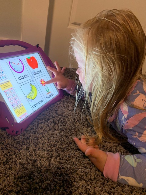 A toddler who is white with blonde hair laying on her stomach on the floor using the tablet; she is pointing to the screen, which shows big symbols and words to teach her language including the words and pictures for “clock”, “apple” and “flower”.