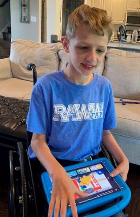 photo of Beckett shows a young blonde-haired boy in a blue t-shirt sitting in his wheelchair in the living room of his house, using his tablet on his lap