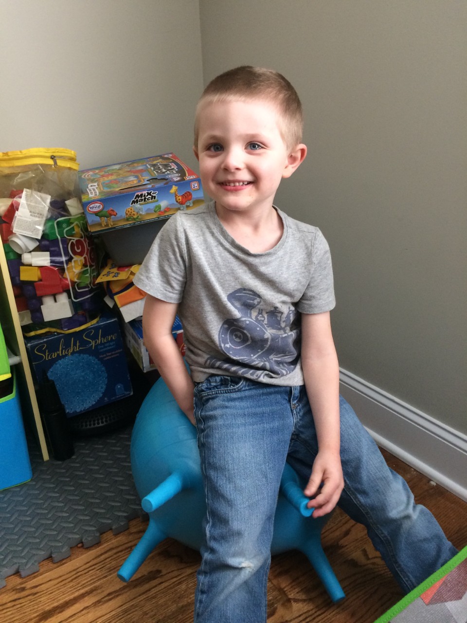 a smiling young blonde boy in a gray t-shirt, sitting on the blue yoga ball with toys and puzzles behind him