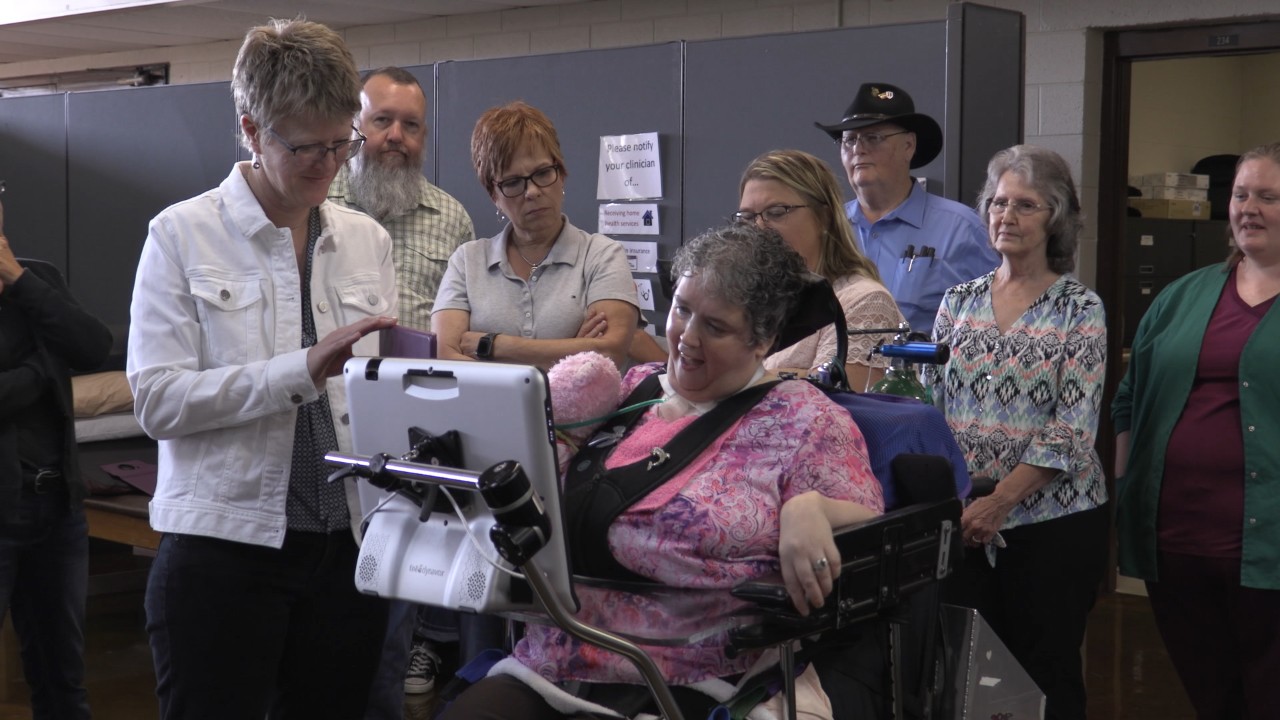 A group of a few adults stand around a middle aged gray haired woman in a power wheelchair who is using a tablet to communicate with them with a text to speech app