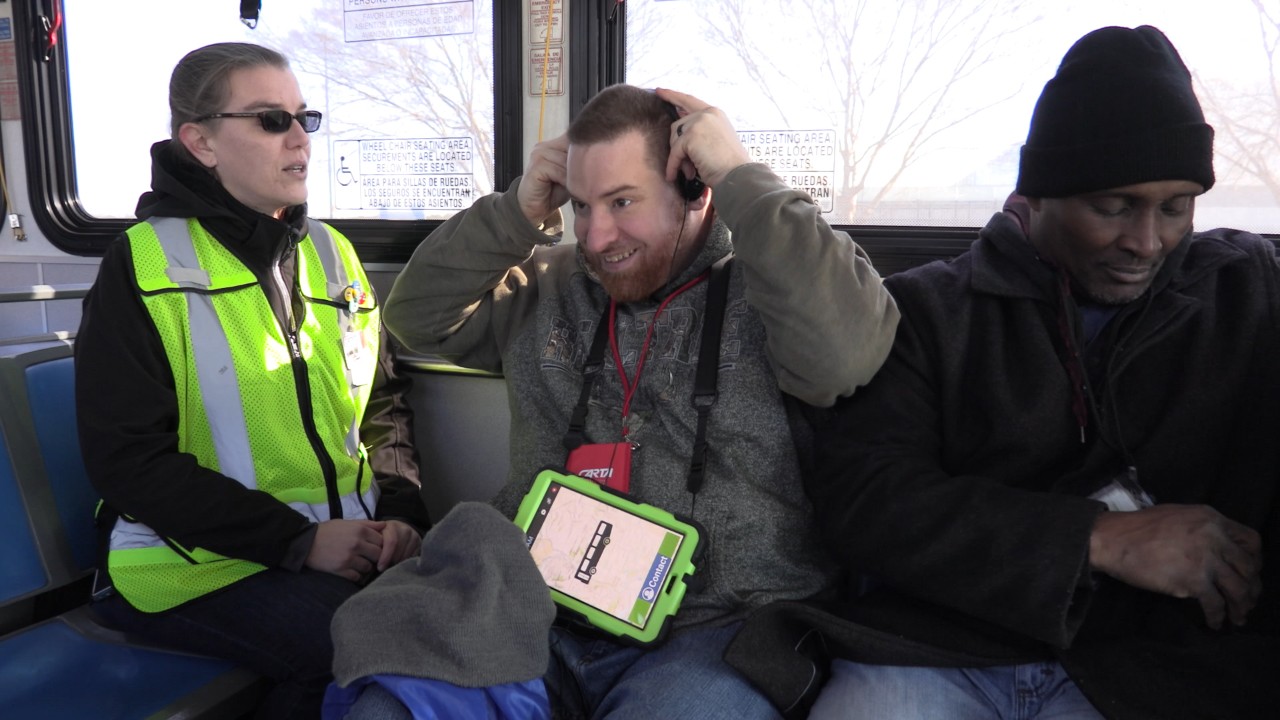 A young man with an intellectual disability is pictured seated on a bus, looking excited, as he puts earphones on and he has a tablet in his lap, attached with a lanyard around his neck to help him communicate