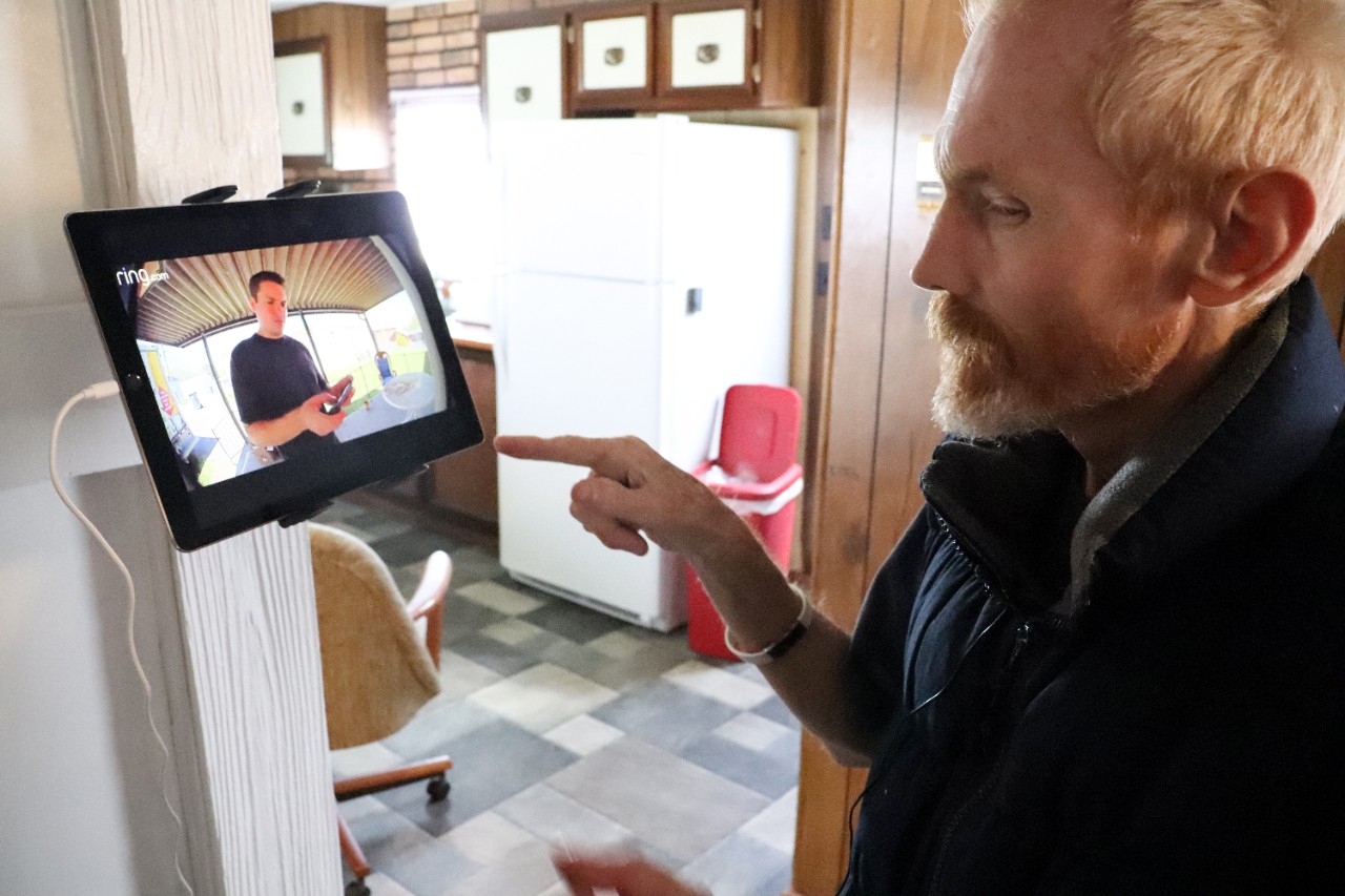 an older white man with a red-blonde hair and beard and sweater in his home kitchen, looking at a tablet mounted on his wall that is showing him who is at his front door (it says the brand name “ring” in the corner of the video on the tablet). His name is Carl and he is about to click the tablet to allow his visitor into his home, after he saw who is at his door