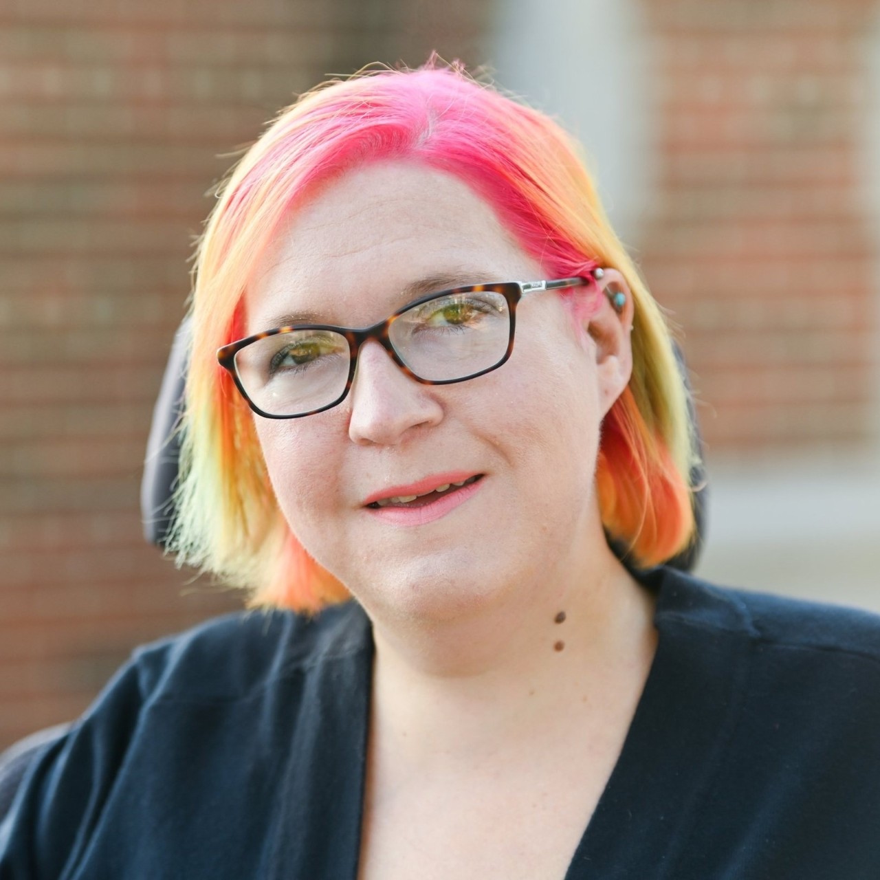 an outdoor headshot of Jean-Marie; she’s sitting in her power wheelchair, and has short straight hair dyed a soft yellow and bright pink; she has a few ear piercings and rimmed glasses and she is smiling