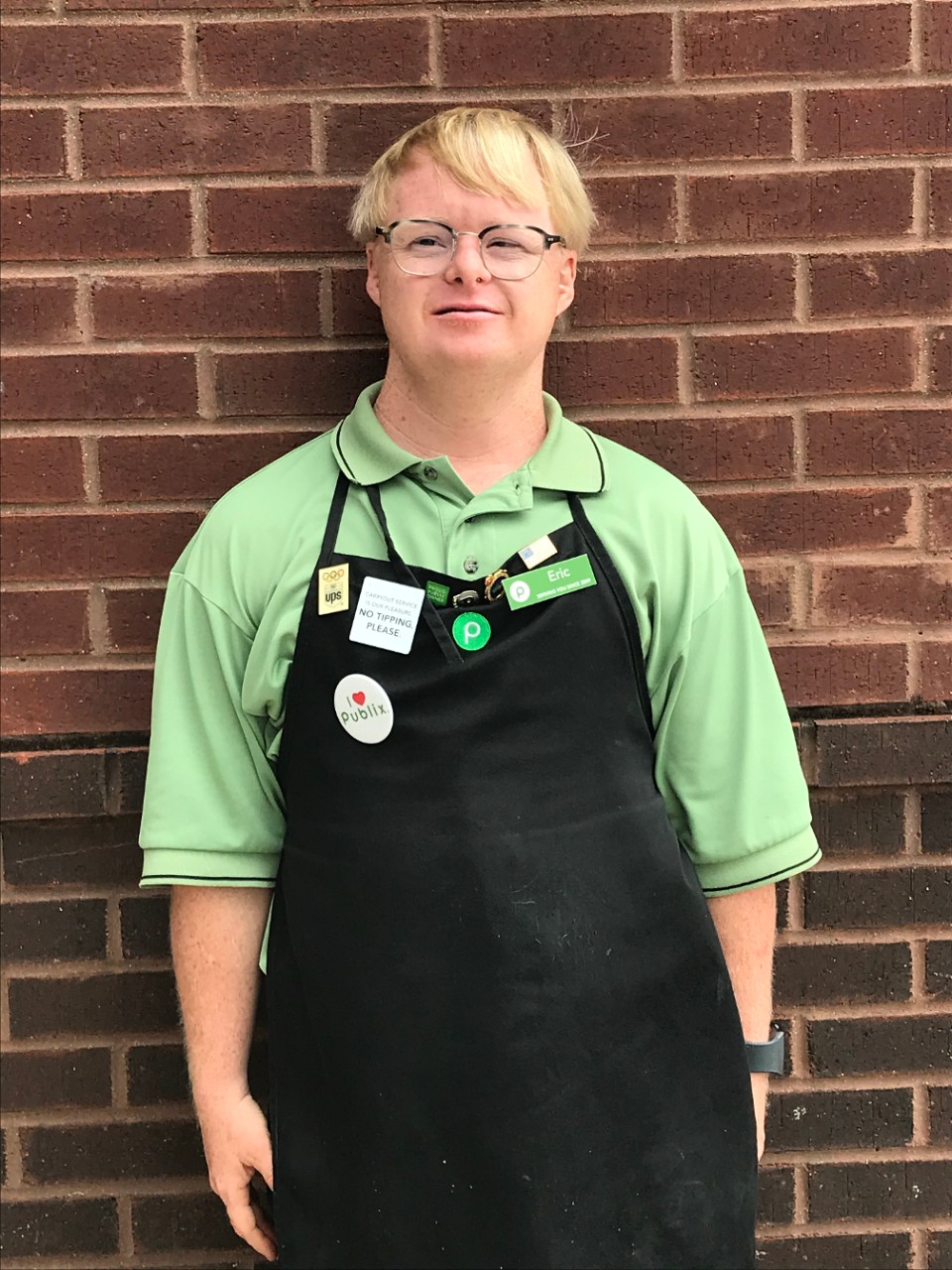 Eric, a young blonde man with Down syndrome and glasses, is wearing his Publix apron and polo shirt standing against a brick wall and smiling