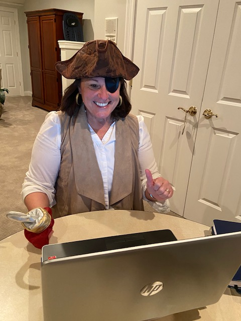 A photo of a woman at her laptop, providing teletherapy to a child and family receiving early intervention services; she is dressed up like a pirate with a pirate hat, vest, eye patch and fake hook on her hand and a big smile on her face to keep the child entertained and engaged in her virtual therapy session.
